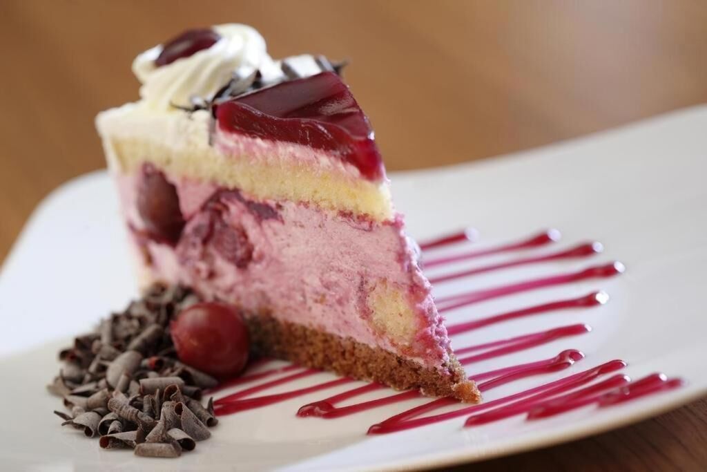 Una porción de postre en capas con cereza encima, servida en un plato blanco con un hilo de salsa roja.