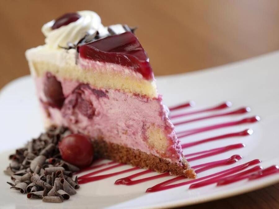 Una porción de postre en capas con cereza encima, servida en un plato blanco con un hilo de salsa roja.