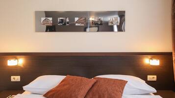 Habitación de hotel con cama, dos almohadas, mesita de noche, lámpara de pared y un collage de fotos enmarcado.
