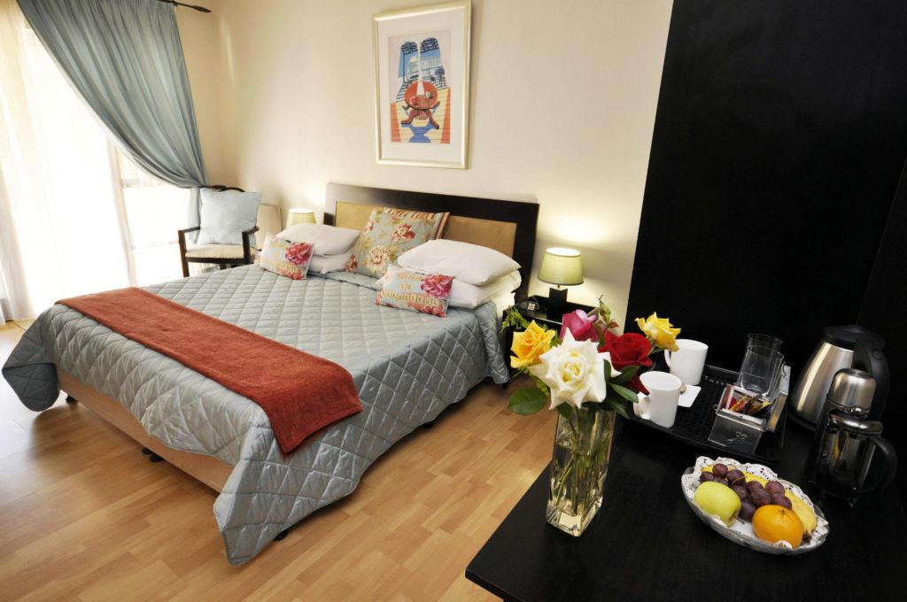 A hotel room with a bed, a bedside table with a vase of flowers, a tray of fruit, and a coffee maker.