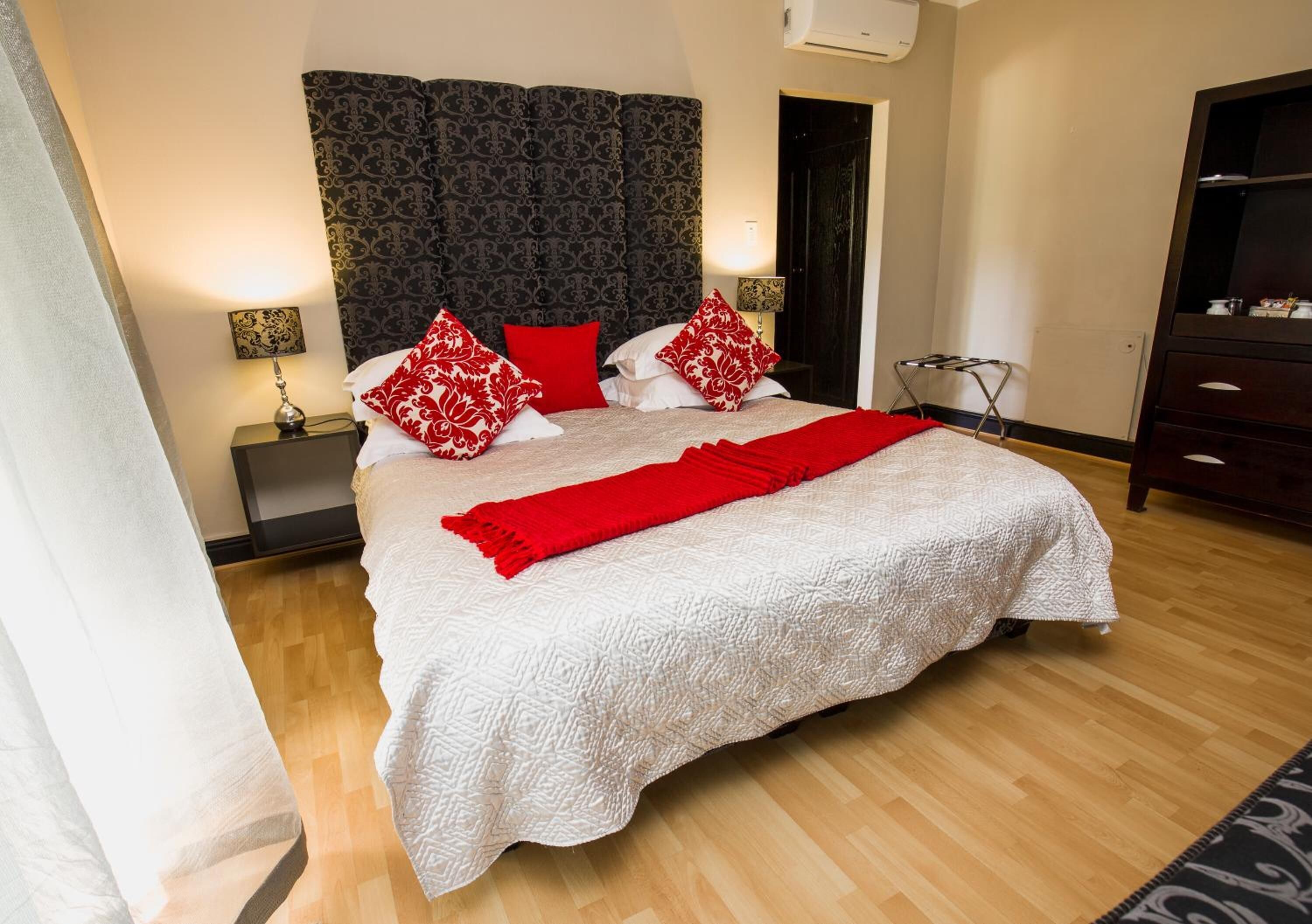 A neatly made bed with a white textured bedspread and red pillows, a dark patterned headboard, and a bedside lamp on a nightstand.