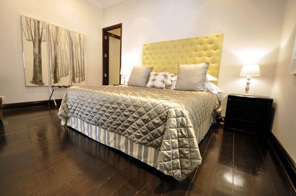 Un dormitorio ordenado con una cama grande, una mesita de noche con lámpara, y una puerta que da a otra habitación.