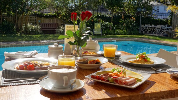 A breakfast setting by a pool with various dishes, a teapot, and a vase with flowers.