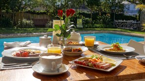 A breakfast setting by a pool with various dishes, a teapot, and a vase with flowers. - Wild Olive Guest House (Cape Town)