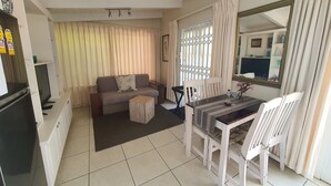 A living room with a sofa, a dining table with chairs, a TV, and a window with curtains.
