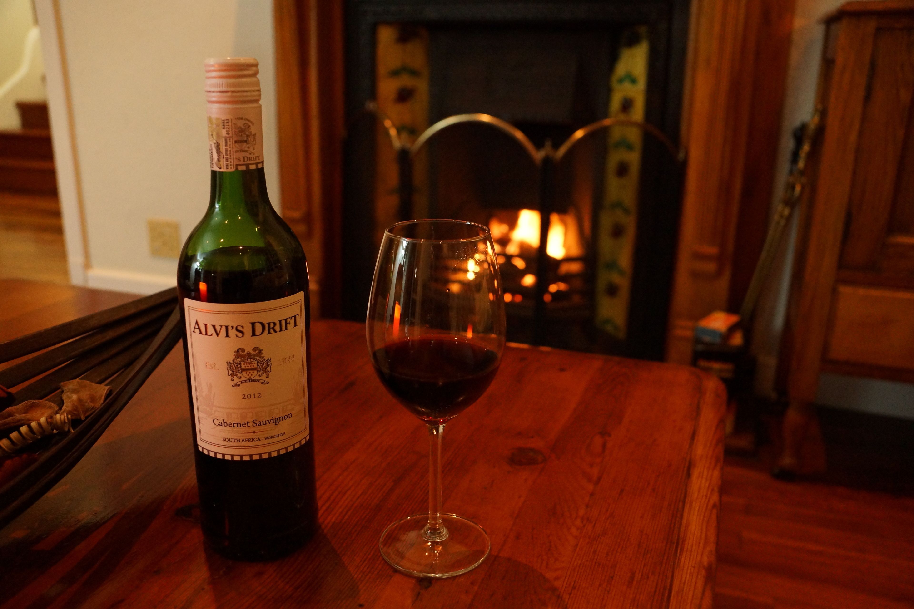 A bottle of Alvi's Drift Cabernet Sauvignon and a glass of red wine on a wooden table in a warmly lit room with a fireplace in the background.