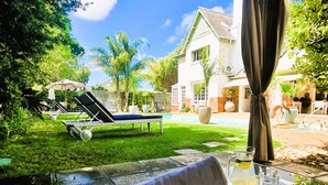 A poolside area with a lounge chair, a table with a towel and glass, and a house in the background.