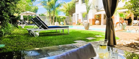Zona junto a la piscina con una tumbona, una mesa con una toalla y un vaso, y una casa al fondo.