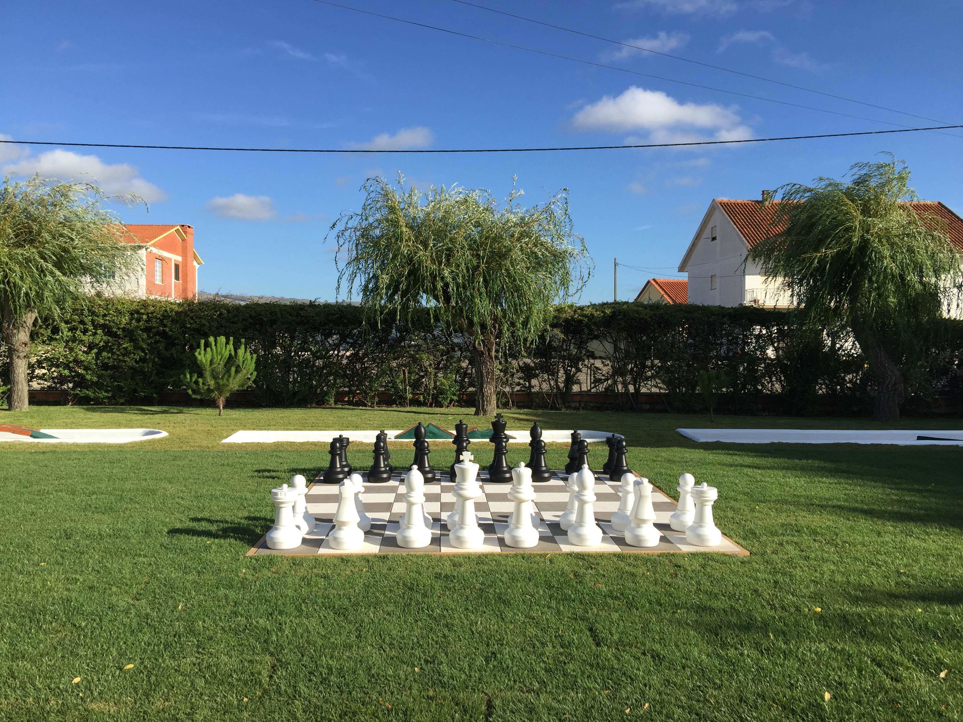 Un tablero de ajedrez dispuesto al aire libre sobre un césped, con árboles y edificios al fondo.