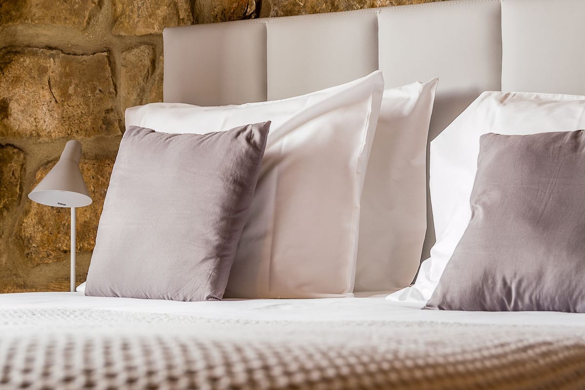 Una cama con almohadas blancas y grises, cabecera blanca y pared de piedra al fondo.