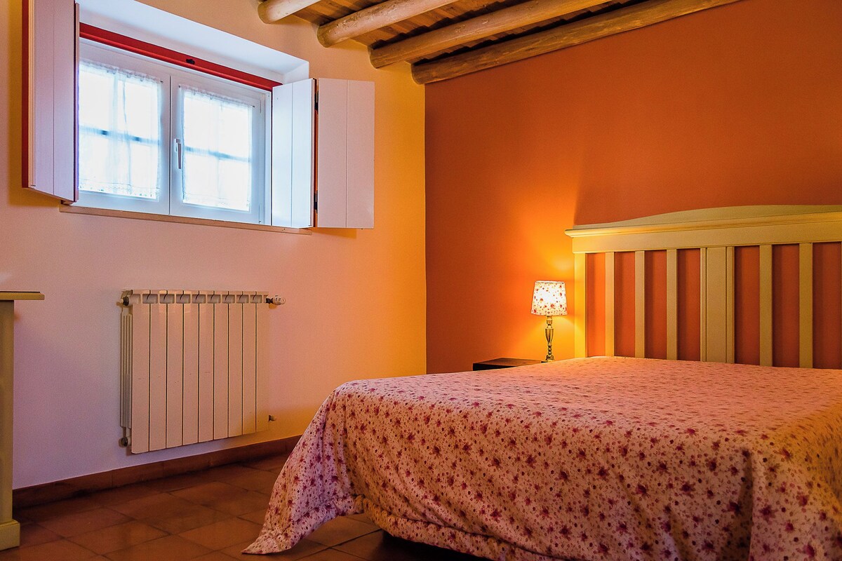 A bedroom with a bed, a window with white shutters, a radiator, and a lamp on a bedside table.
