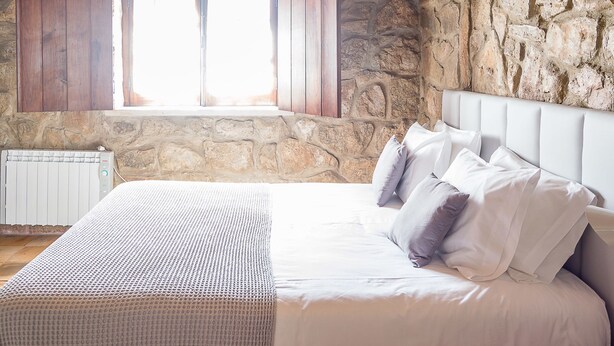 A bedroom with a stone wall, a window with wooden shutters, a white bed with multiple pillows, and a radiator. - Casa da Padeira - TER (Alcobaça)