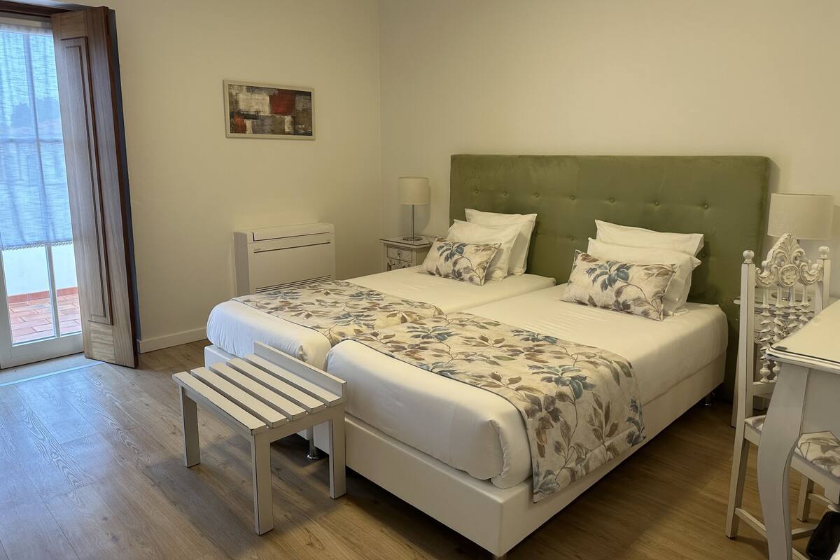 Un dormitorio ordenado con cama, mesitas de noche, un banco y una ventana.