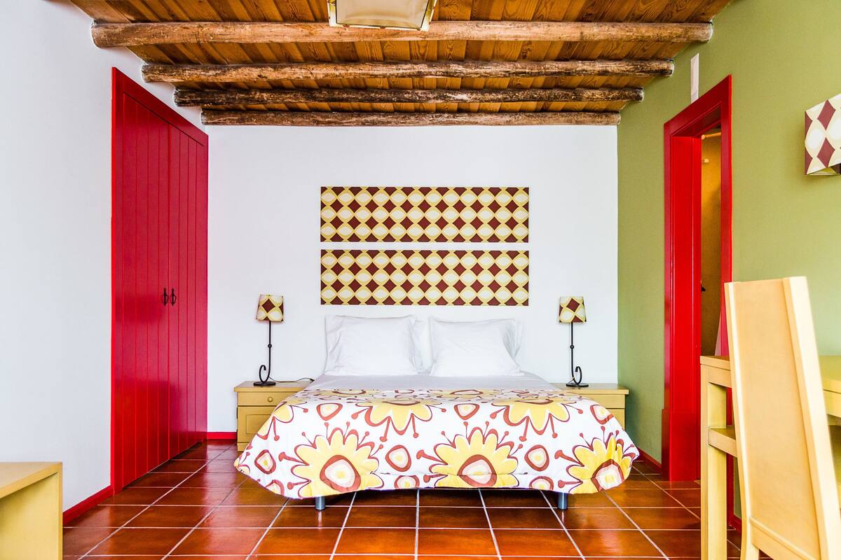 Un dormitorio con cama, dos mesitas de noche, una puerta roja, un cabecero estampado y techo de madera.