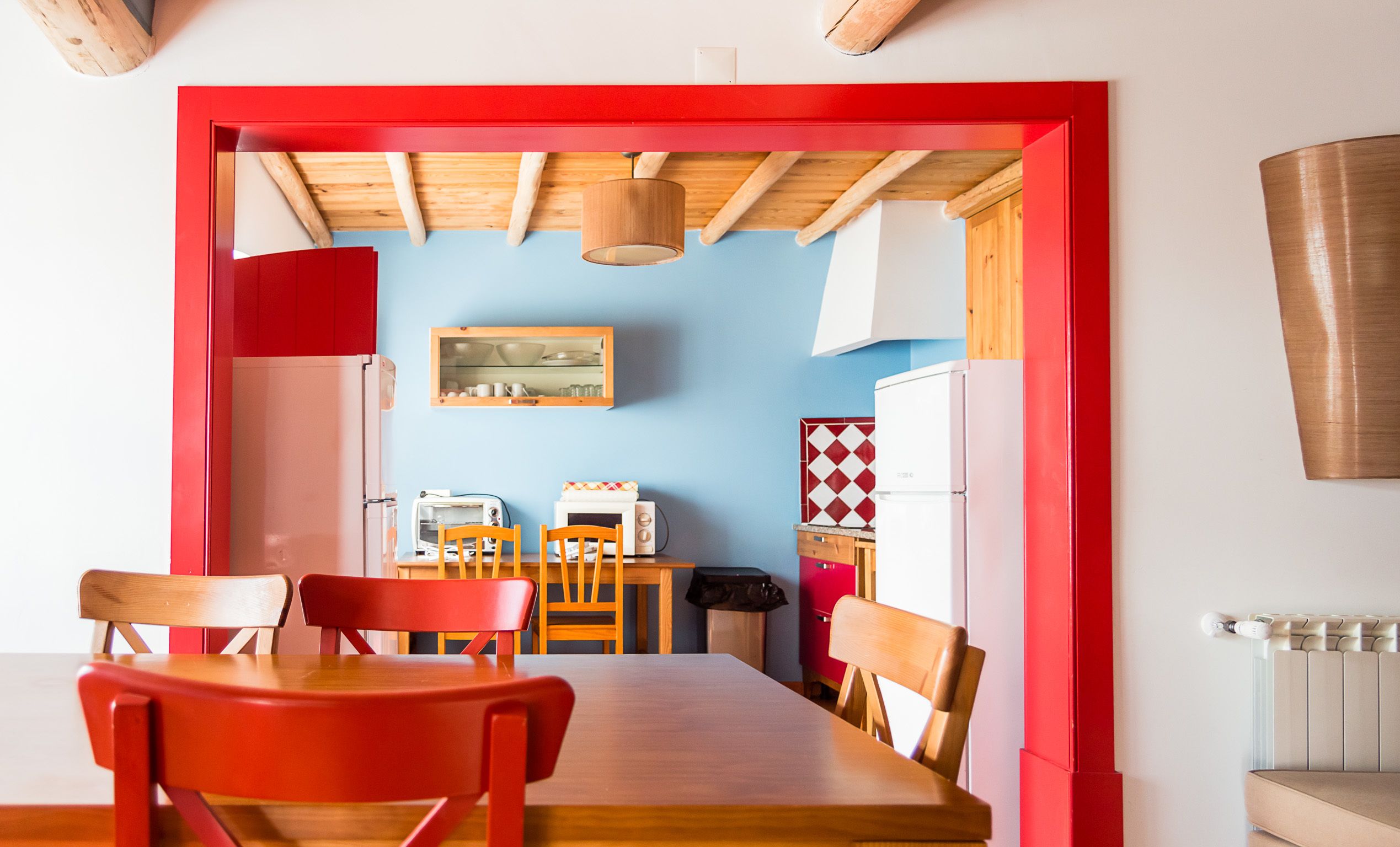 Un comedor con una mesa de madera y sillas rojas, una cocina con microondas, tostadora y armarios, y una pared azul con un cuadro enmarcado.