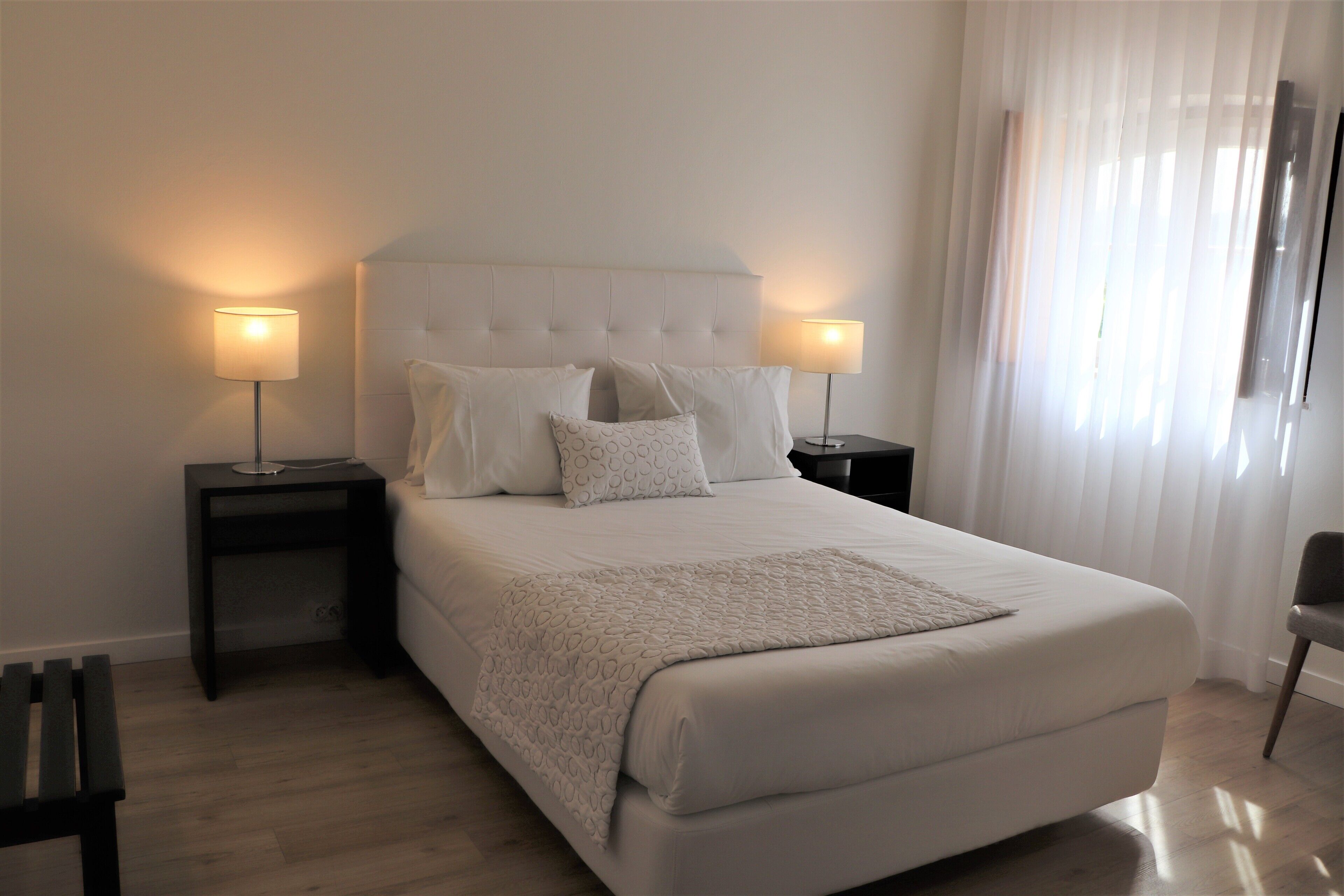 Una cama bien tendida con cabecera, dos mesitas de noche con lámparas, una silla y un ventanal con cortinas traslúcidas.