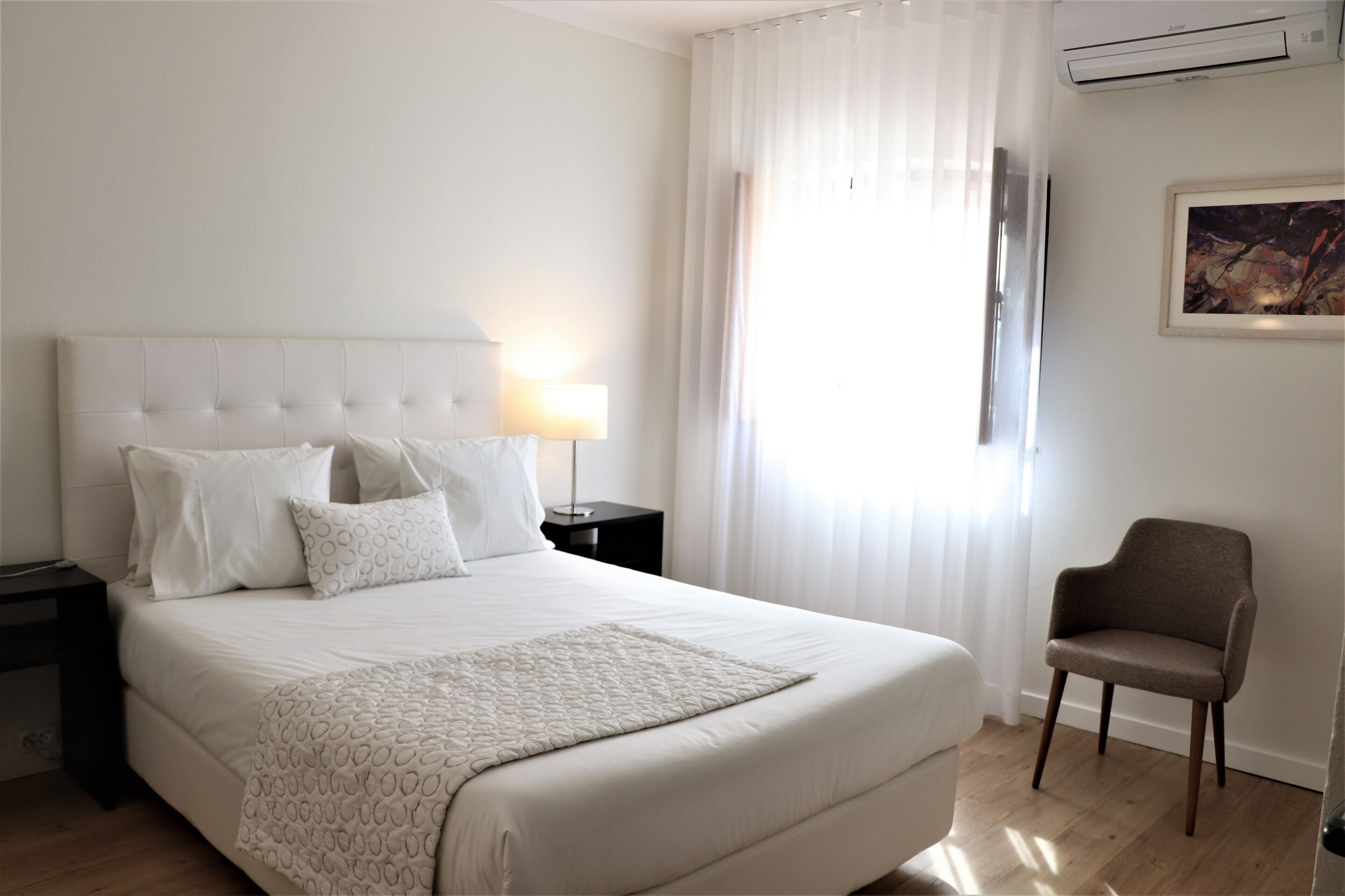 Ein Hotelzimmer mit einem großen Bett, einem Nachttisch, einem Sessel und einem Fenster mit Vorhängen.