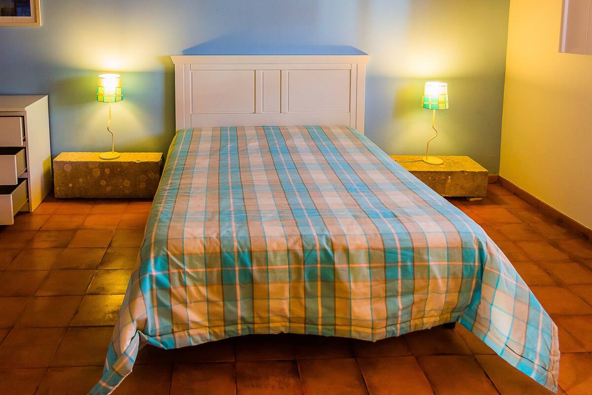 Una cama con colcha escocesa, dos lámparas de noche, un cabecero blanco y suelo de madera.