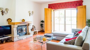 A living room with a fireplace, a sofa, a television, and a large window with curtains. - Casa da Padeira - TER (Alcobaça)