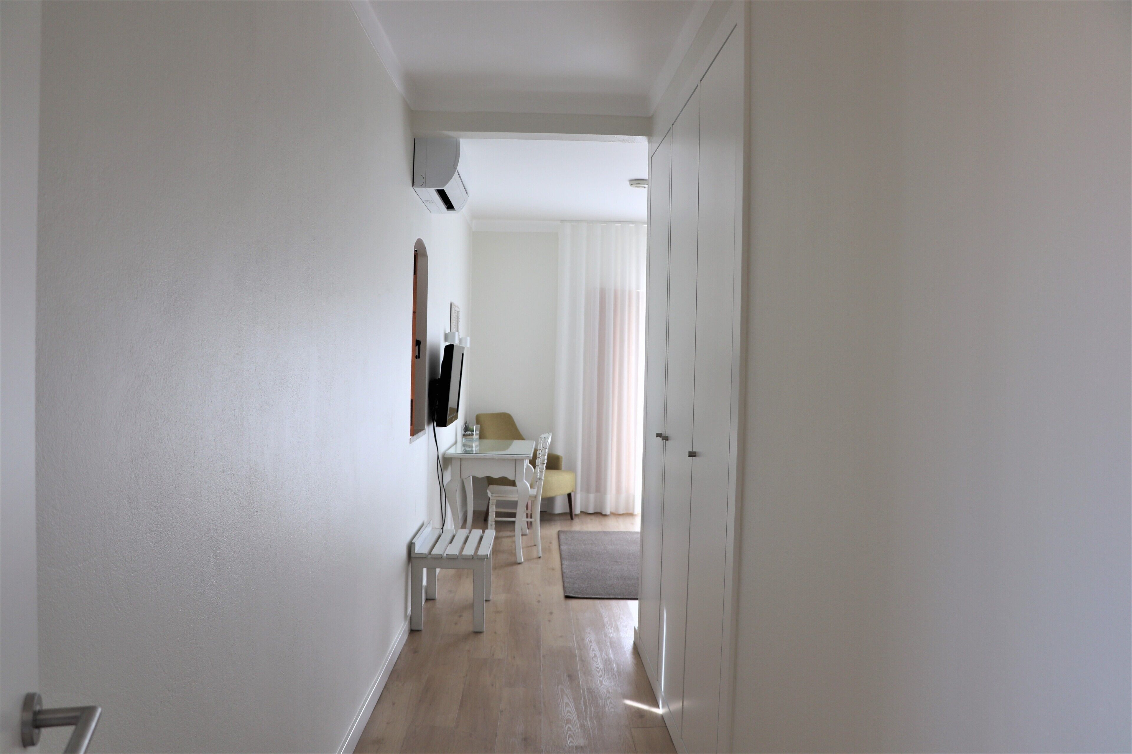 A hallway with a white bench, a table with chairs, and a TV mounted on the wall.