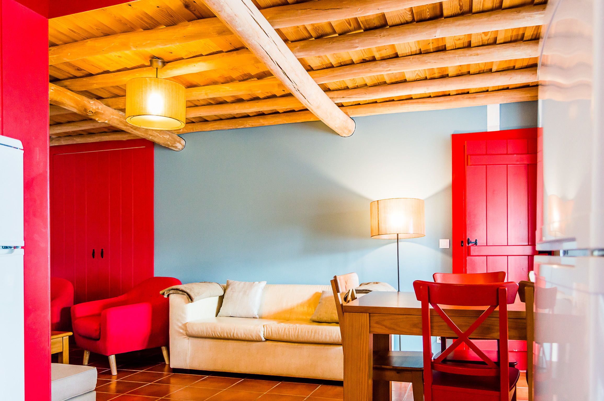 Una habitación con techo de madera, una puerta roja, un sofá color crema y una silla roja, junto con una mesa de madera.
