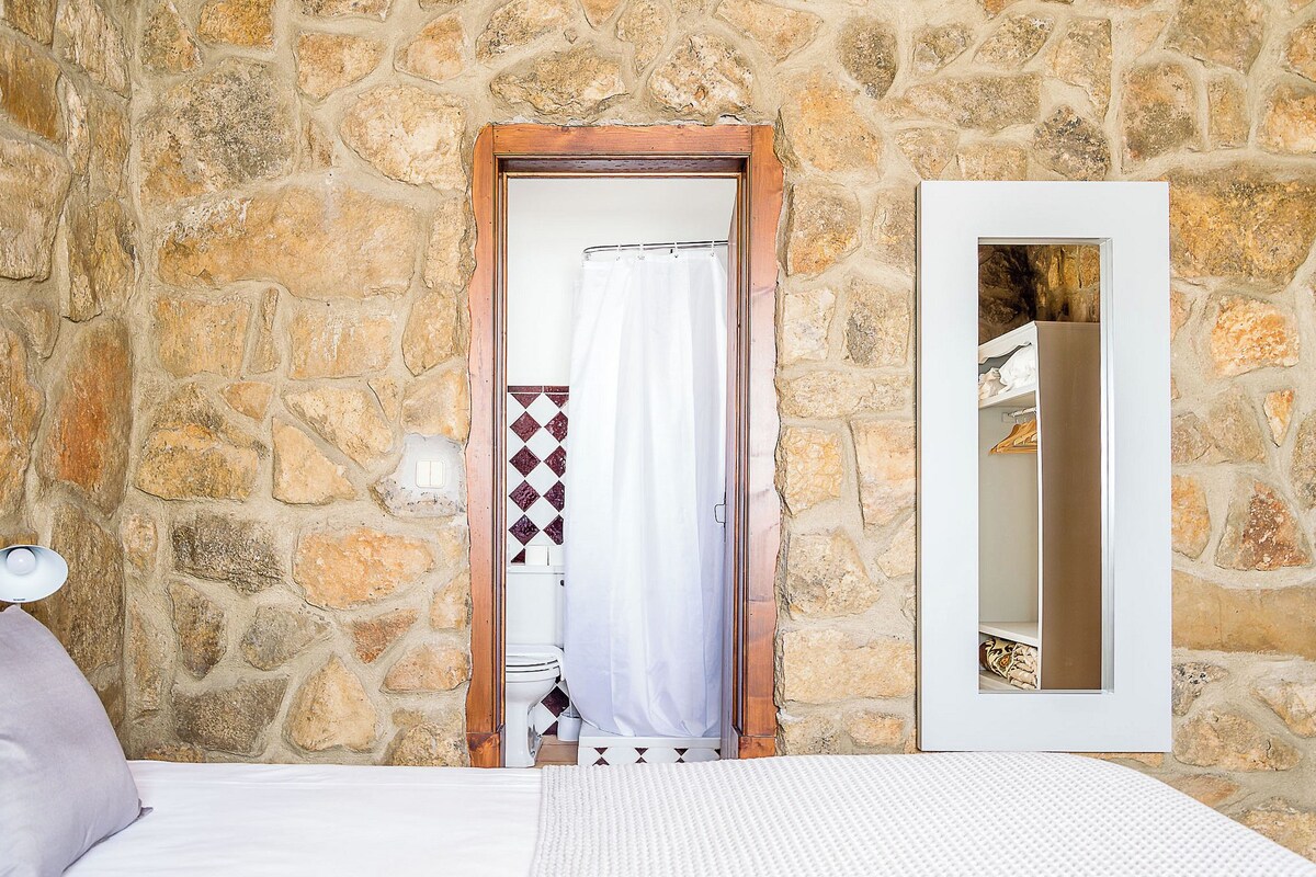 A bedroom with a stone wall, a wooden door, a white bed, and a mirror.