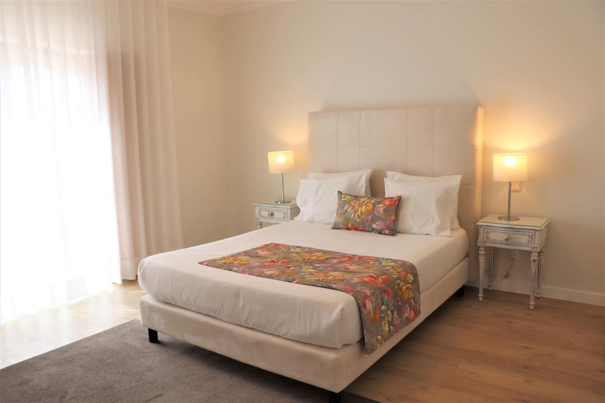 Una cama bien tendida con una colcha estampada, dos mesitas de noche con lámparas y una ventana con cortinas traslúcidas.