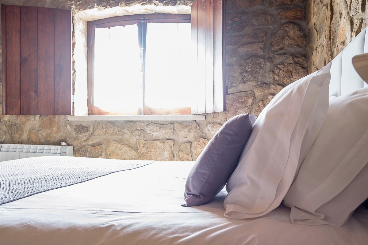 A bedroom with a stone wall, a window with wooden shutters, and a bed with white bedding and a gray throw blanket.