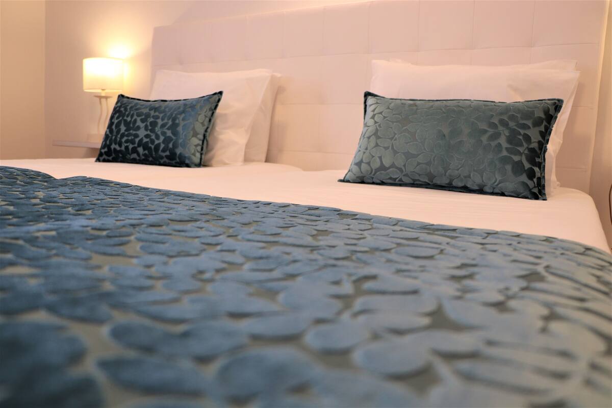 Una cama bien hecha con una colcha estampada y dos almohadas, una lámpara de noche y un cabecero blanco.