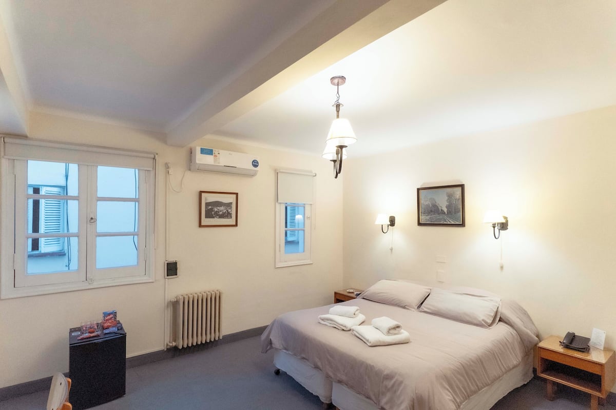 Habitación de hotel con cama, mesitas de noche, radiador, ventana y aire acondicionado instalado en la pared.