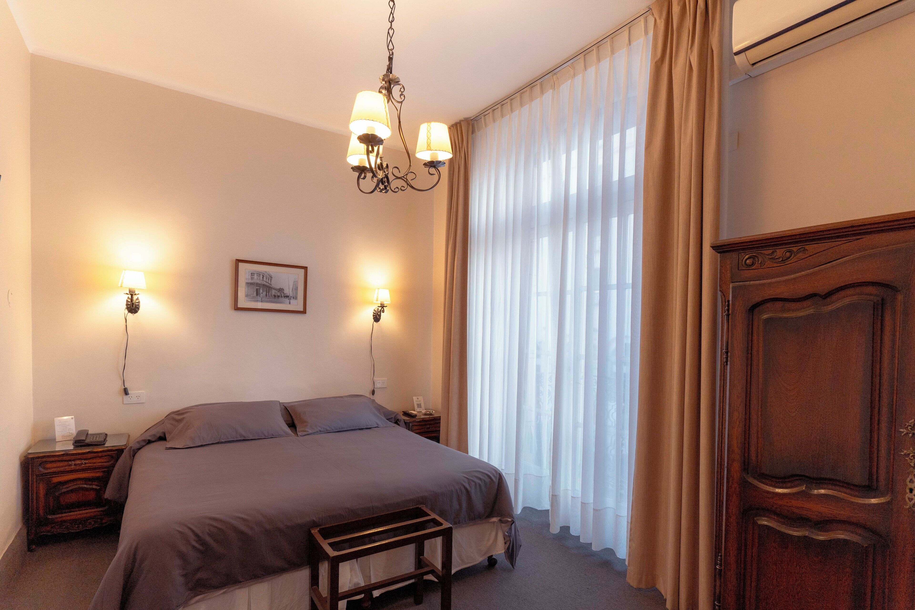 A bedroom with a large bed, two bedside lamps, a wooden nightstand, a wooden wardrobe, and a chandelier.
