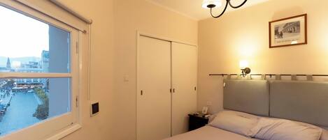 A hotel room with a bed, a nightstand, a lamp, a framed picture on the wall, and a window with a city view.