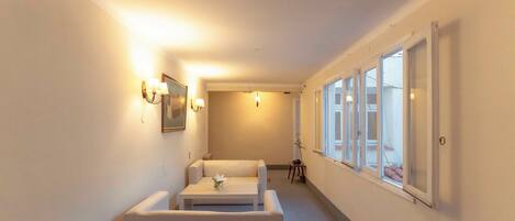 A narrow hallway with a sofa and a small table, a framed picture on the wall, and a window with blinds.