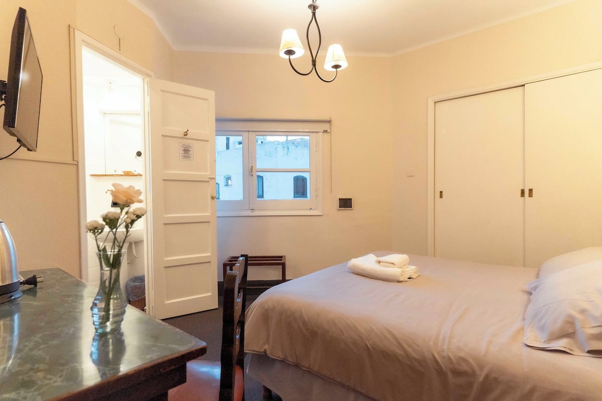 Habitación de hotel con cama, escritorio con un jarrón de flores, una silla, ventana y candelabro.