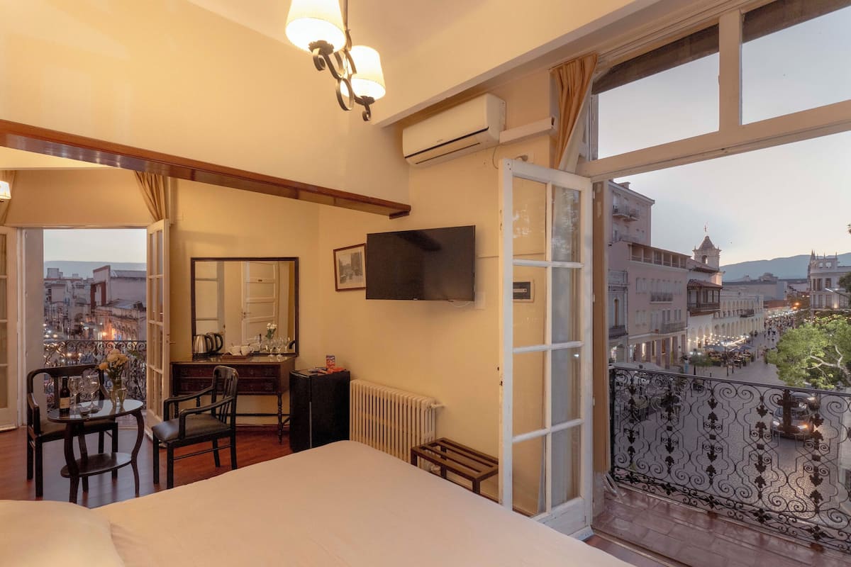 Habitación de hotel con balcón, cama, escritorio con silla, televisor y vista panorámica de la ciudad.