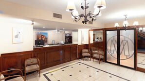 A hotel lobby with wooden reception desk, chairs, a chandelier, and a large mirror with a logo.