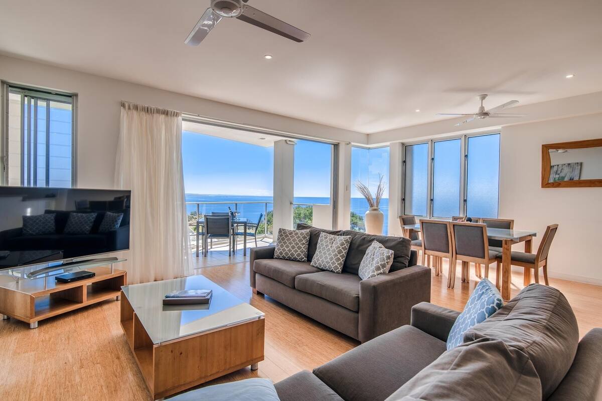 Amplia sala de estar con televisor grande, asientos cómodos, comedor y balcón con vista al mar.