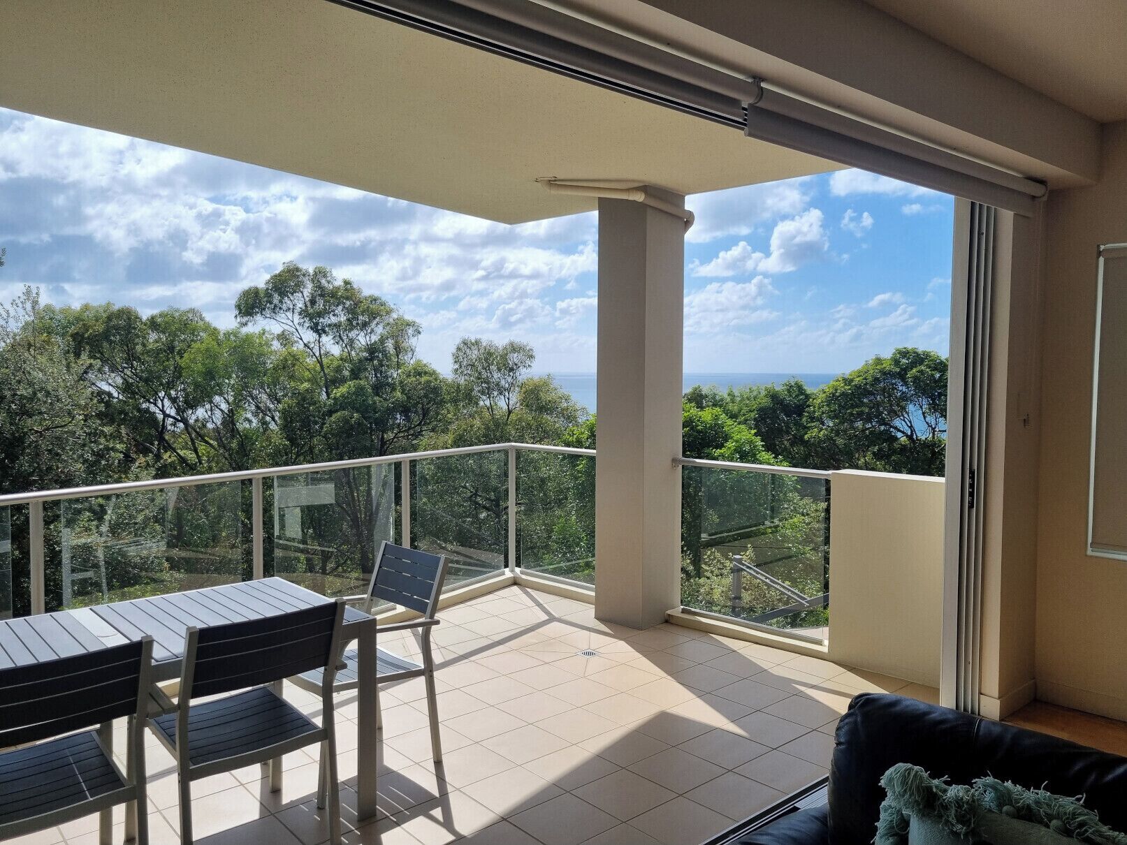 A balcony with a table and chairs, a glass railing, and a view of trees and a distant landscape.