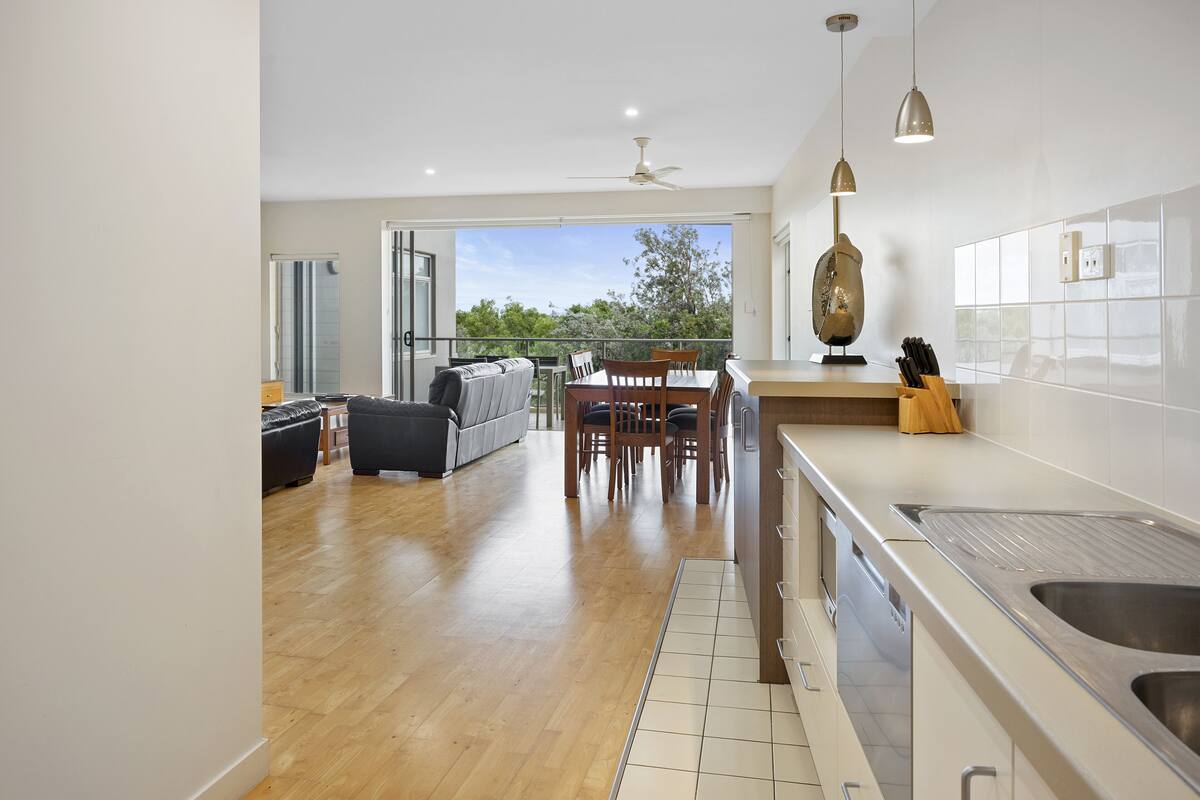 Una cocina moderna con una isla grande, una zona de comedor con mesa y sillas, y un balcón con vistas a árboles.