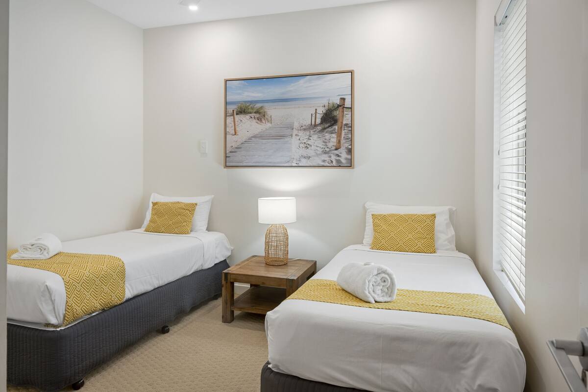 Habitación de hotel con dos camas, un cuadro de un sendero en la playa, una lámpara y una ventana con persianas.