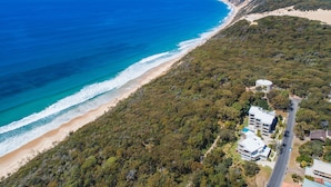 Vista aérea de una zona costera con playa, bosque frondoso y una carretera que lleva a un edificio de varios pisos y piscina.