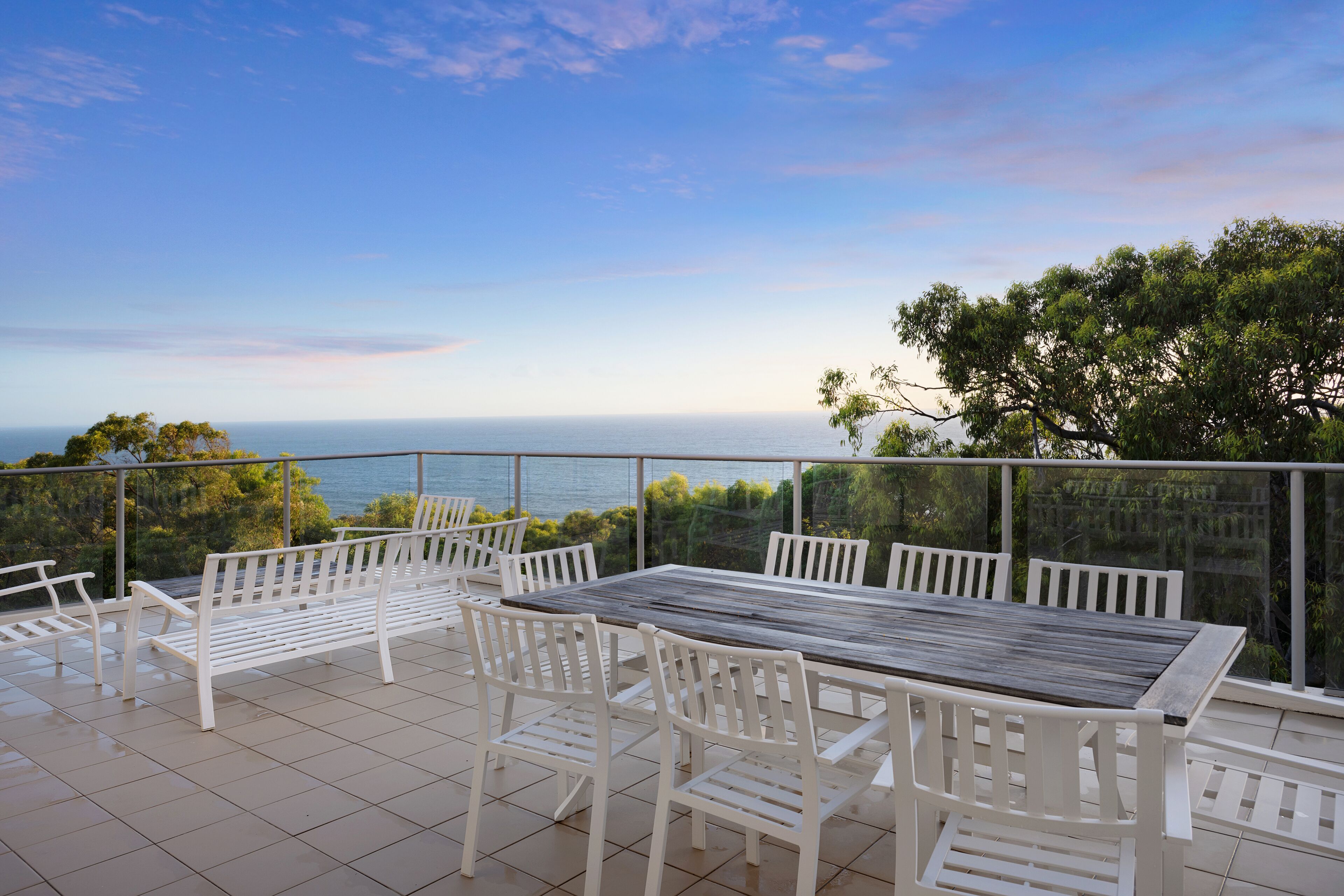Une terrasse avec une table en bois et des chaises blanches, offrant une vue panoramique sur la mer et les arbres.