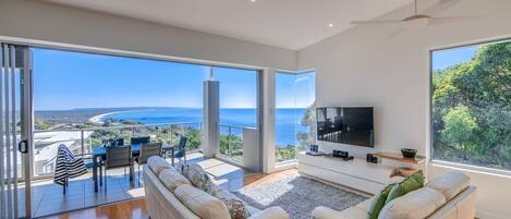 A spacious living room with a large TV, comfortable seating, and a balcony with a view of the sea.