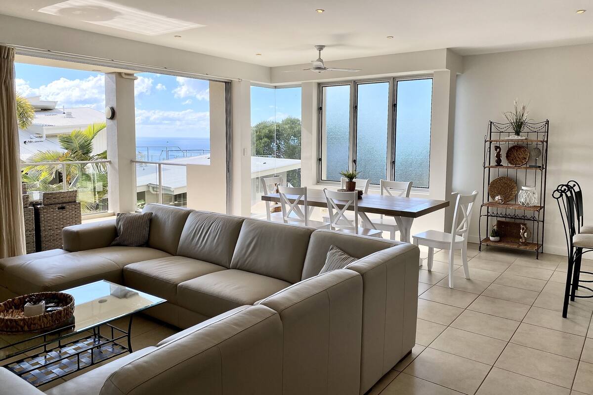 Una sala de estar moderna con un sofá beige, una mesa de centro de vidrio y una zona de comedor con sillas blancas y una mesa de madera. Amplias ventanas ofrecen vistas al mar y al verdor.