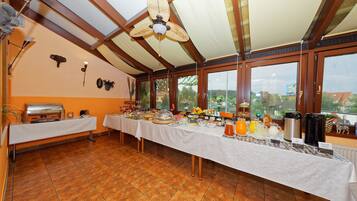 A buffet setup with a variety of food and drink options, a ceiling fan, and large windows offering a view of the outdoors.