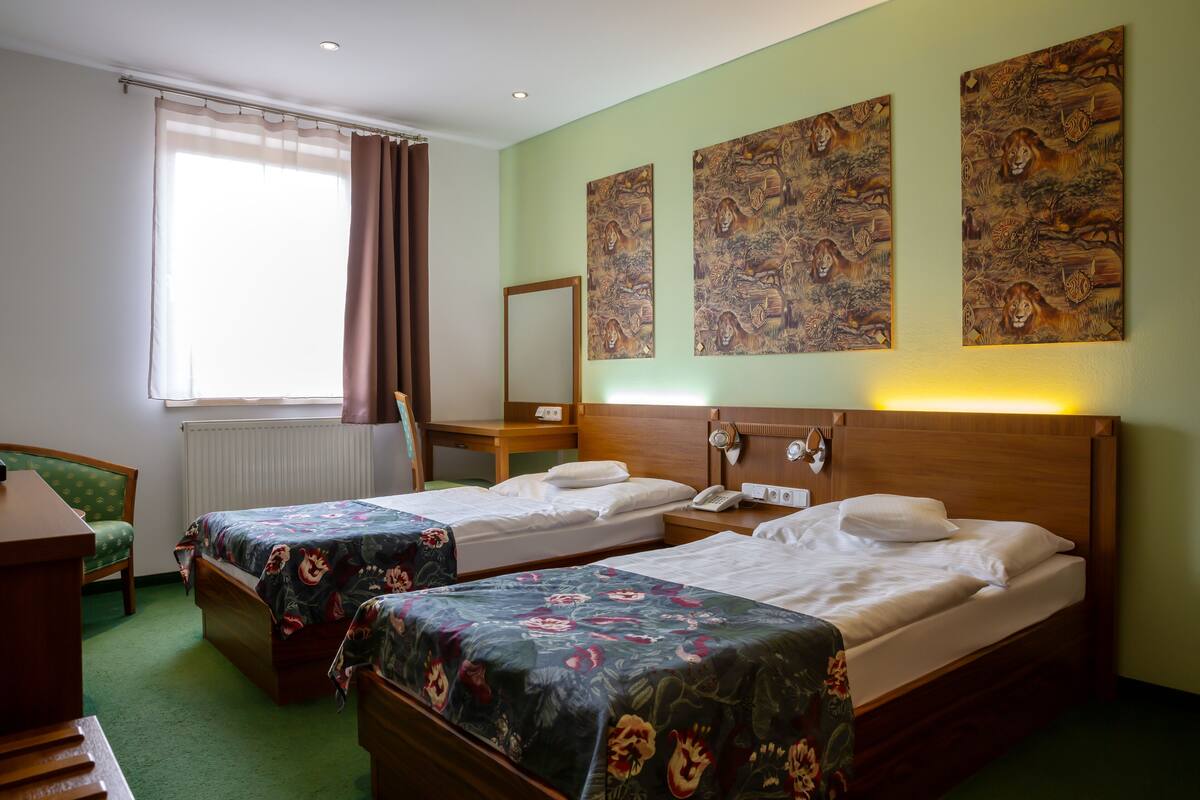 Quarto de hotel com duas camas de solteiro, uma escrivaninha de madeira, uma cadeira, um radiador e obras de arte nas paredes.