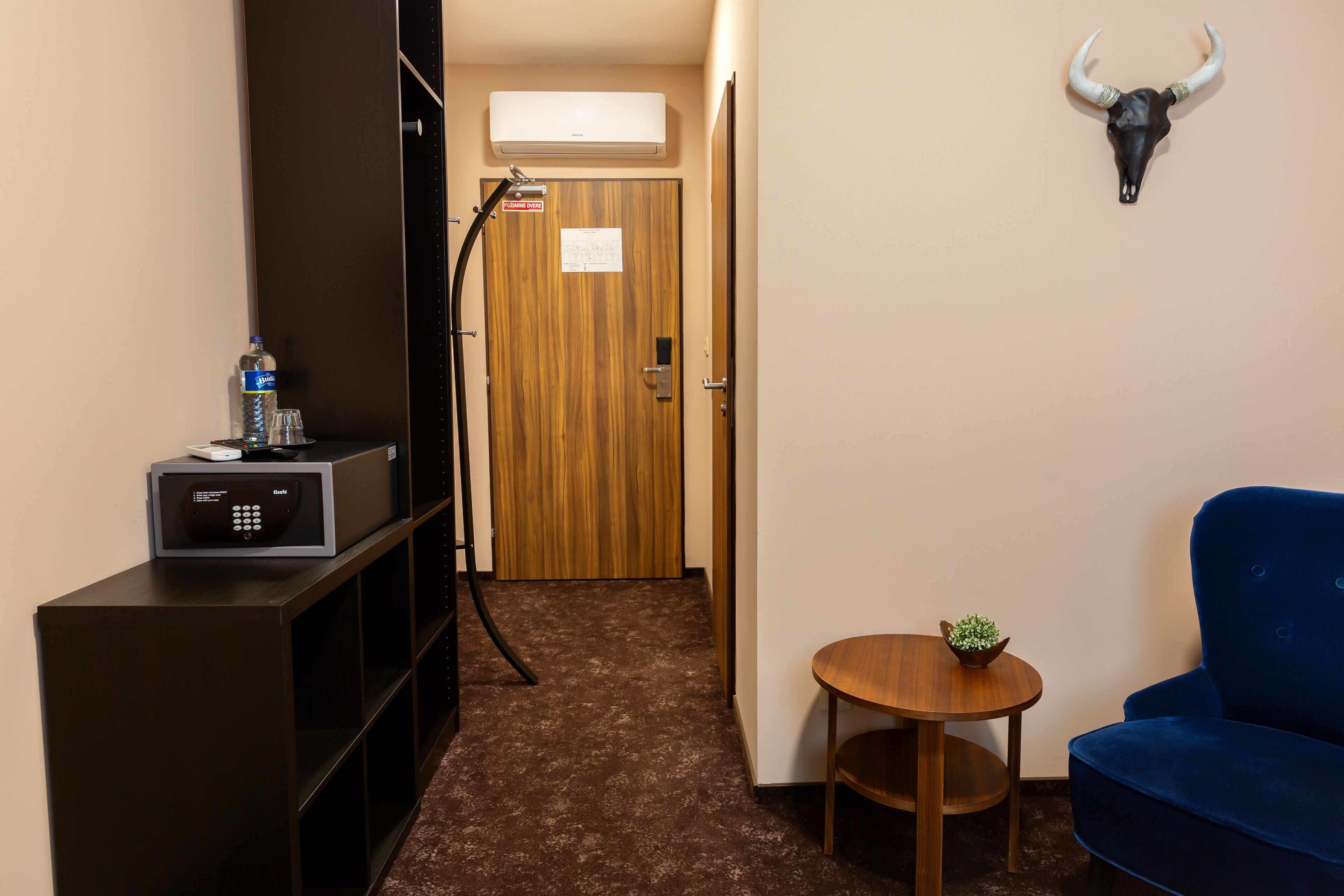 Een hotelkamer met een houten deur, een blauwe bank, een klein houten tafeltje met een plant, een minibar en een op de muur bevestigde decoratie van een stierenhoofd.