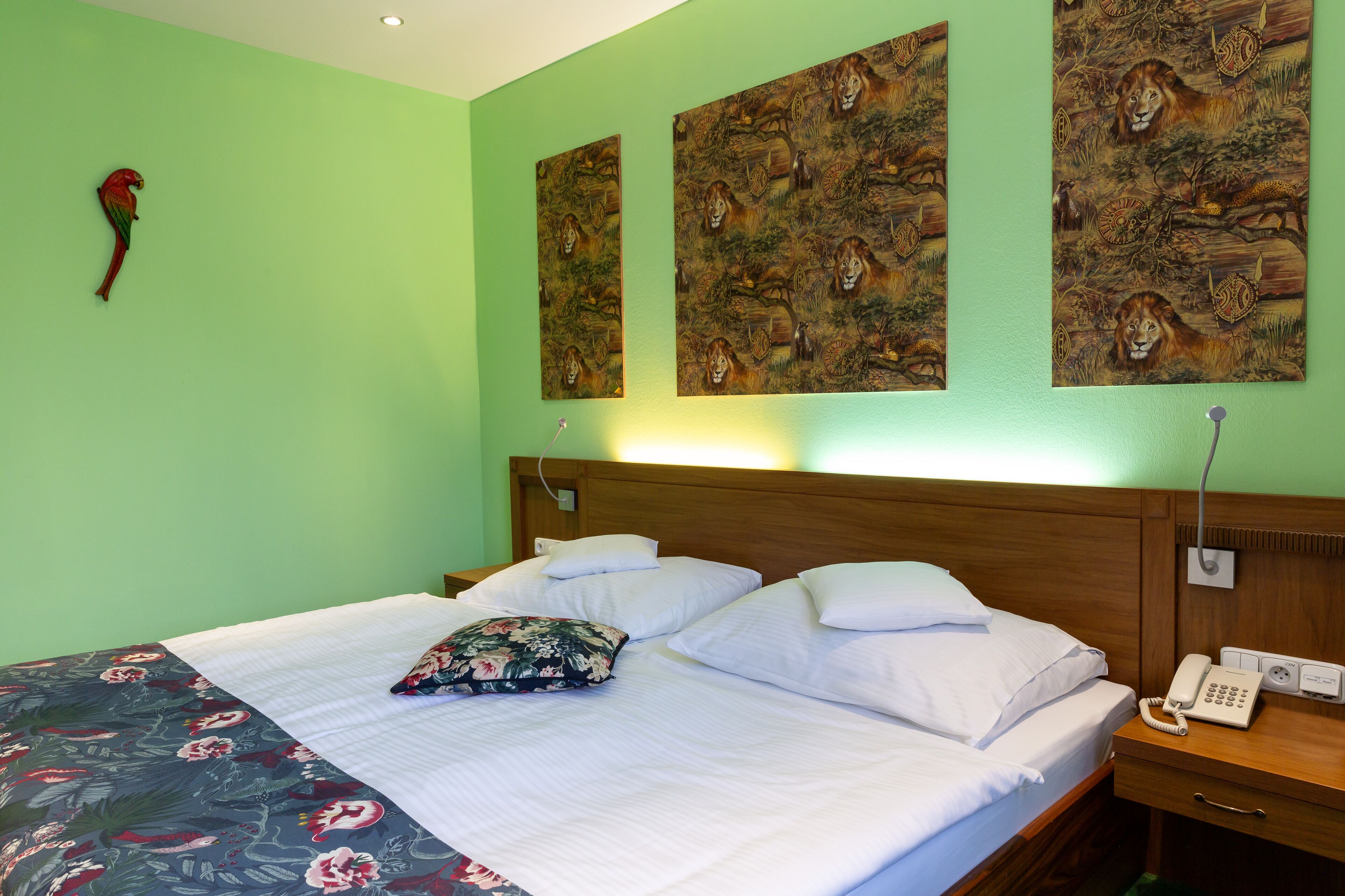 Habitación de hotel con pared verde, cabecero de madera, cama con ropa blanca, manta estampada con motivos florales y un teléfono sobre una mesita de noche de madera.