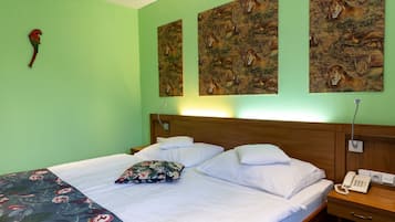 Habitación de hotel con pared verde, cabecero de madera, cama con ropa blanca, manta estampada con motivos florales y un teléfono sobre una mesita de noche de madera.