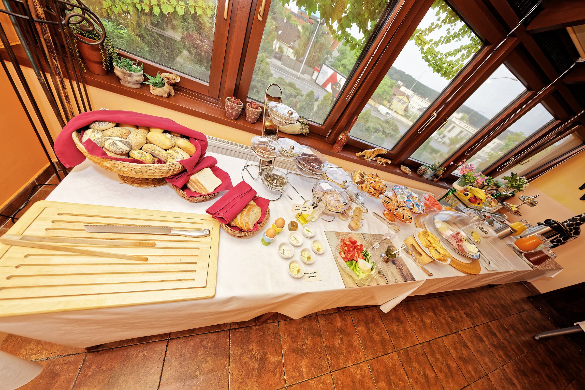 A buffet table with various food items and utensils, set against a window with a view of buildings and greenery.
